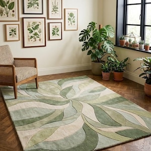 May include: A rectangular area rug featuring a large, swirling floral-inspired pattern in various shades of sage green, olive, and cream. The design consists of overlapping, curved petal-like shapes that radiate from a central point across the surface.