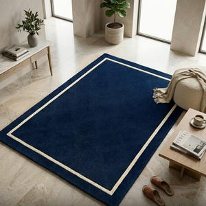May include: A rectangular area rug featuring a deep navy blue center field enclosed by a thick, cream-colored border. The rug has a solid, uniform texture and is positioned flat on a light-colored stone floor in a bright, open interior space.
