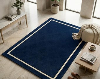 May include: A rectangular area rug featuring a deep navy blue center field enclosed by a thick, cream-colored border. The rug has a solid, uniform texture and is positioned flat on a light-colored stone floor in a bright, open interior space.