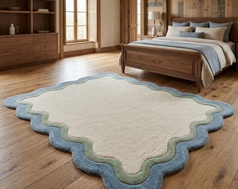 May include: A rectangular area rug featuring a cream-colored center with scalloped edges. The rug is bordered by two distinct wavy bands, one in a soft sage green and the outermost layer in a muted blue, creating a layered, rippled effect.