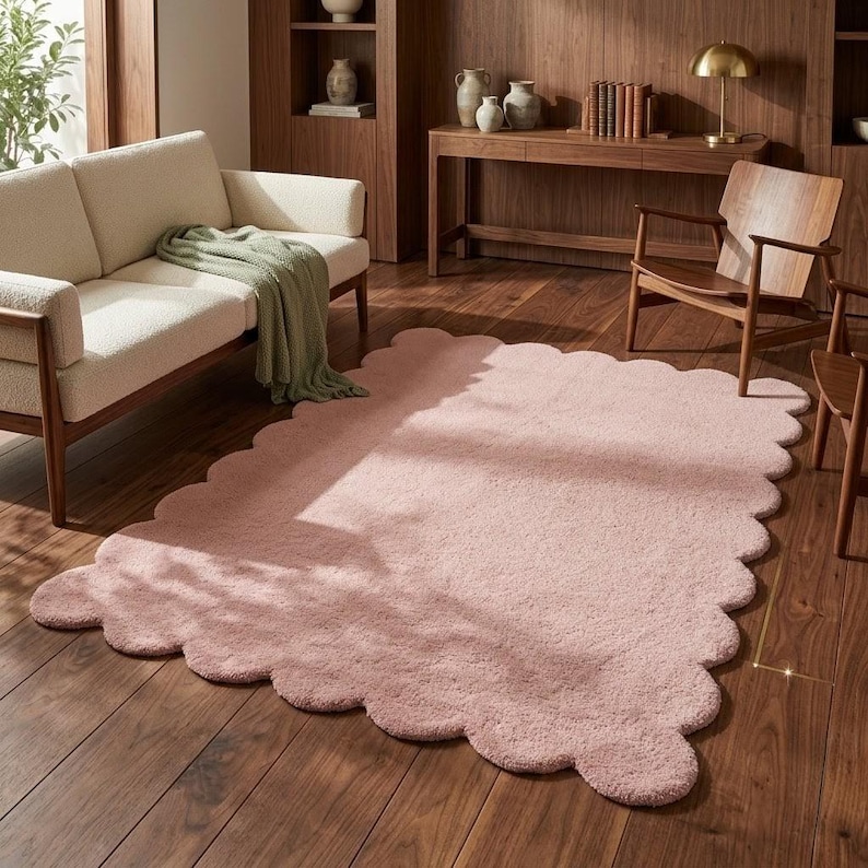 Scalloped edge pink area rug featuring a soft plush texture and playful wavy border design. This cloud-inspired floor carpet adds a cozy decorative accent to nurseries, bedrooms, living rooms, and reading corners. The gentle pink color complements cream furniture, pastel accessories, light wood finishes, and Scandinavian-inspired interiors. Its rounded edges create a soft, whimsical appearance that works beautifully in modern homes seeking warmth, comfort, and understated decorative charm.