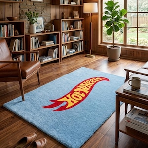 May include: A rectangular light blue area rug featuring the Hot Wheels logo in yellow and red centered on the surface. The rug has a soft, textured pile and is positioned on a wooden floor in a room with bookshelves and a leather chair.