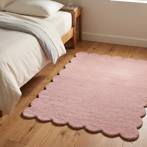 Pink wavy border area rug with a rounded scalloped outline and cozy plush styling. This decorative carpet brings a playful yet refined touch to bedrooms, living rooms, nurseries, and reading spaces. Its soft color coordinates easily with beige furniture, ivory bedding, pale wooden accents, pastel decor, and minimalist accessories. The cloud-like silhouette makes it a distinctive floor accent for Scandinavian, modern, contemporary, and soft eclectic interiors.