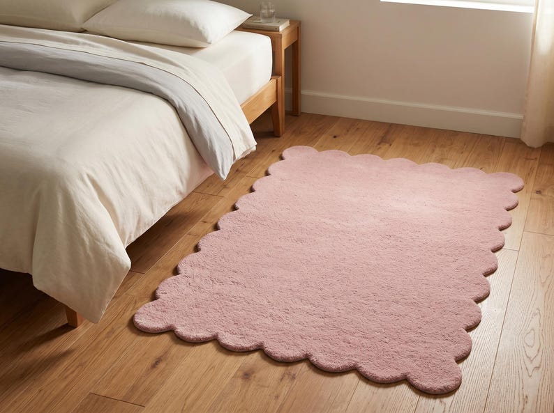 Pink wavy border area rug with a rounded scalloped outline and cozy plush styling. This decorative carpet brings a playful yet refined touch to bedrooms, living rooms, nurseries, and reading spaces. Its soft color coordinates easily with beige furniture, ivory bedding, pale wooden accents, pastel decor, and minimalist accessories. The cloud-like silhouette makes it a distinctive floor accent for Scandinavian, modern, contemporary, and soft eclectic interiors.