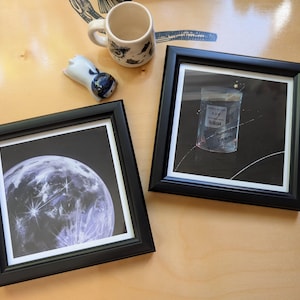 May include: Two square black frames containing art prints. One print depicts a glowing, textured moon against a dark background. The other print shows a small, clear plastic bag labeled 'THUNDER FARM' floating in space with stars and orbital lines.