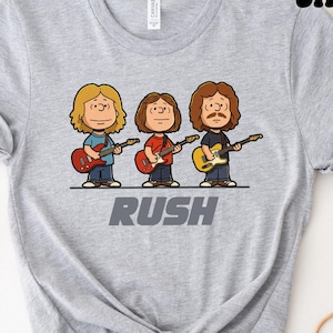 Op de afbeelding: Een grijs gemêleerd T-shirt met korte mouwen en een cartoonafbeelding van drie muzikanten die gitaar spelen. Onder de figuren staat het woord RUSH in dikke, grijze hoofdletters. Het T-shirt heeft een ronde hals en een standaard pasvorm.