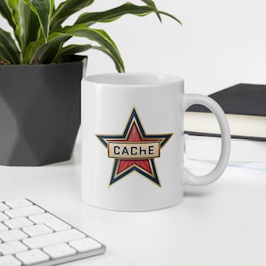 Puede incluir: Una taza de cerámica blanca con un gráfico de una estrella roja con un borde dorado y azul oscuro. En el centro de la estrella hay una banda horizontal con fondo dorado y borde azul oscuro, que muestra la palabra CACHE en letras azules.