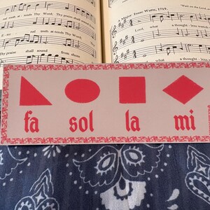 Peut inclure: Un marque-page rectangulaire avec un fond beige clair et une bordure décorative rouge. Il présente quatre formes géométriques rouges—un triangle, un ovale, un carré et un losange—au-dessus du texte rouge fa, sol, la et mi. Le texte hell.md.scissors est à droite.