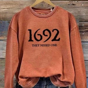 May include: A rust-colored crewneck sweatshirt featuring the black text 1692 and the phrase THEY MISSED ONE printed on the front. The garment has long sleeves, a ribbed neckline, and a slightly distressed, washed-out fabric appearance.