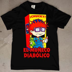 Puede incluir: Una camiseta negra de manga corta con un gráfico de un personaje de dibujos animados con pelo rojo y gafas moradas, vistiendo un peto azul y sosteniendo un juguete de dinosaurio verde y un biberón. El texto dice ¡CHUCKY! y EL MUÑECO DIABÓLICO.