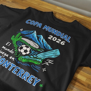 May include: A black short-sleeve t-shirt featuring a graphic print with blue mountains, a soccer ball, a green cactus, and a soccer cleat. The text reads COPA MUNDIAL 2026 and El mundo juega en MONTERREY in blue and white lettering with small stars.