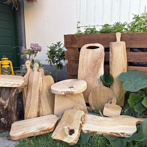 Può includere: Una collezione di taglieri in legno chiaro e un piccolo sgabello in legno con bordi naturali e venature del legno visibili. Gli articoli sono disposti in varie forme e dimensioni, inclusi design rettangolari, ovali e rotondi, che mettono in risalto la consistenza grezza del legno.