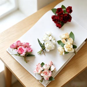 Puede incluir: Cinco prendidos de rosas artificiales dispuestos sobre una superficie blanca. El conjunto incluye un ramillete de rosas rojo oscuro, blanco, crema, rosa intenso y rosa claro, cada uno con hojas de tela verde y atado con cuerda de color marrón.