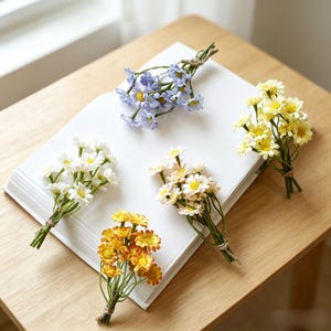 Puede incluir: Cinco pequeños ramos de flores artificiales tipo margarita en blanco, azul claro, amarillo, naranja y rosa pálido, cada uno atado con un cordel fino. Los tallos son verdes y cada ramo presenta varias flores pequeñas con centros amarillos.