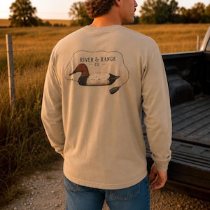May include: A long-sleeve beige t-shirt featuring a graphic on the back of a duck decoy with a line and weight. The text RIVER & RANGE CO. is printed above the duck illustration. The shirt is worn with blue jeans and a tan baseball cap.
