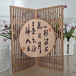 May include: A folding wooden screen with a vertical slat design and a central circular beige panel featuring black Chinese calligraphy. The screen is positioned upright, showcasing a light brown wood grain finish and intricate cutouts.