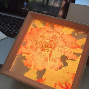 May include: A square light box featuring a warm, illuminated display of a large, detailed orange and yellow peony flower illustration. The artwork is encased in a light-toned wooden frame, providing a soft glow that highlights the intricate petal patterns and organic shapes of the floral design.