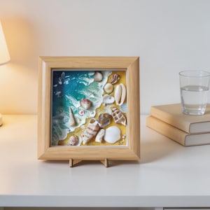 Könnte beinhalten: Ein quadratischer Holzrahmen mit einem Kunstwerk aus Harz, das eine Strandszene mit einem Farbverlauf von blauem Meerwasser und gelbem Sand darstellt. Verschiedene weiße und braune Muscheln sind entlang der Küstenlinie im Harz eingebettet.
