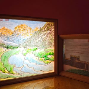May include: Two square wooden frames containing illuminated landscape artwork. The larger frame displays a vibrant painting of terraced rice fields and mountains. The smaller frame features a painting of a dark structure against a pale sky.