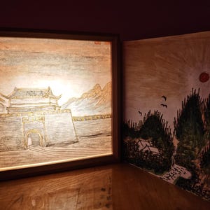 May include: A square, illuminated wooden frame containing a drawing of a traditional gatehouse with mountains in the background, positioned next to a flat, square panel featuring a painted landscape with a red sun, green trees, and birds.