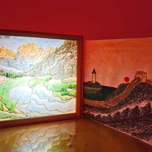 May include: A square light box with a wooden frame displays a painting of terraced fields and mountains. Beside it stands a square print featuring a painting of a tower, a red sun, and a section of the Great Wall of China against a warm sky.