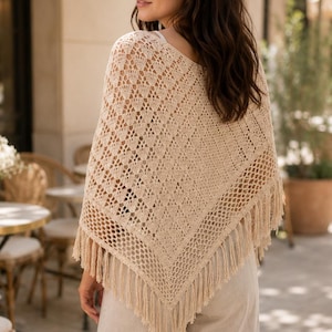 Puede incluir: Un poncho de ganchillo beige con silueta triangular, intrincados patrones calados y largos flecos en los bordes inferiores. La prenda se lleva sobre pantalones claros, mostrando una construcción de punto ligera y texturizada.