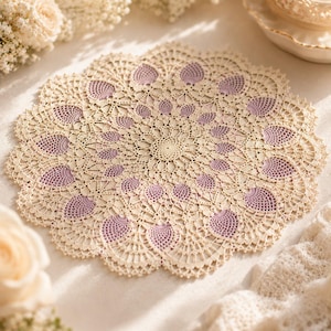 May include: A round, intricate crochet doily featuring a cream-colored thread base with repeating, petal-like motifs filled with soft lavender-colored thread. The design radiates from a central point, showcasing delicate, open-work lace patterns.