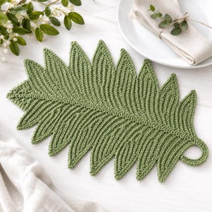 May include: A sage green crochet placemat shaped like a large fern leaf with a textured, ribbed surface and a small circular loop at the base for hanging. The placemat features a detailed, symmetrical vein pattern throughout the entire leaf design.