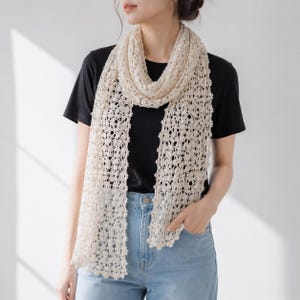 May include: A cream-colored, open-work crochet scarf featuring a delicate, repeating floral-inspired pattern. The scarf is draped around the neck, hanging down in two long, vertical panels with scalloped edges, showcasing a fine, lightweight texture.