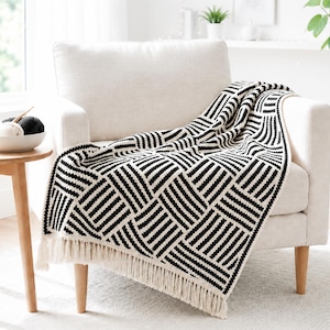 Op de afbeelding: Een rechthoekige plaid met een contrastrijk zwart-wit geometrisch patroon van herhalende gestreepte vierkanten. Het textiel is afgewerkt met lange, crèmekleurige franjes langs de onderrand, wat zorgt voor een getextureerd en decoratief uiterlijk.