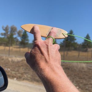Op de afbeelding: Een klein vinger-surfplankje van lichtgekleurd hout met een gestructureerde grip-pad op de bovenzijde. Een dun, felgroen touwtje is bevestigd aan de achterkant van het plankje en een bijpassende groene touwlus zit om de middelvinger.