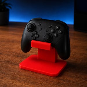 May include: A black wireless video game controller resting on a bright red plastic stand. The controller features two analog sticks, a directional pad, and four action buttons labeled A, B, X, and Y, along with central menu and capture buttons.