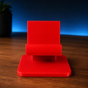 May include: A bright red mobile phone stand featuring a flat square base and a vertical back support with a horizontal ledge designed to hold a device in place. The stand has a smooth, matte finish and a simple, functional geometric construction.
