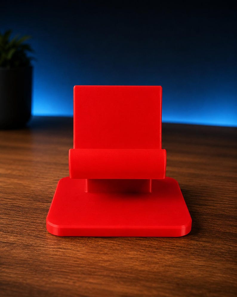 May include: A bright red mobile phone stand featuring a flat square base and a vertical back support with a horizontal ledge designed to hold a device in place. The stand has a smooth, matte finish and a simple, functional geometric construction.