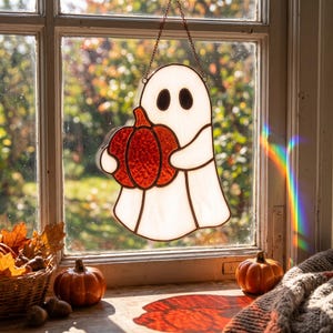 May include: A stained glass suncatcher shaped like a white ghost holding a textured orange pumpkin. The ghost has two black oval eyes and is suspended by a thin metal chain. The piece features dark lead lines outlining the ghost and pumpkin.