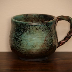 May include: A ceramic mug with a rounded body featuring a mottled teal and dark brown glaze. The surface has a textured, horizontal finish. It includes a distinctive handle shaped like a twisted rope with dark green and brown tones.