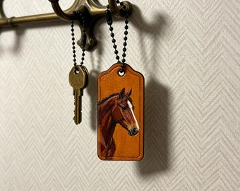 May include: A rectangular wooden keychain featuring a hand-painted portrait of a brown horse with a white blaze on its face. The tag hangs from a black ball chain, accompanied by a brass-colored metal key on a separate black ball chain.