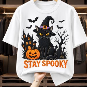 Può includere: Una t-shirt bianca a maniche corte con una grafica raffigurante un gatto nero con un cappello da strega, una zucca, una casa stregata, pipistrelli e un albero spoglio. Il testo "STAY SPOOKY" è stampato in lettere arancioni sul fondo.