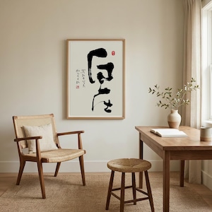 May include: A framed wall art print featuring bold black Japanese calligraphy on a light cream background. The artwork includes smaller vertical text and a small red circular seal. The print is displayed in a light wood frame on a plain wall.