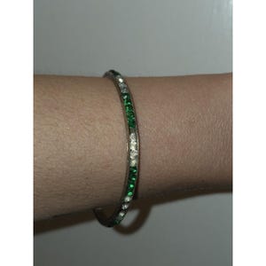 May include: A thin, circular metal bangle bracelet featuring an alternating pattern of small, round, clear white stones and vibrant green stones set along the entire outer surface of the band, creating a continuous, sparkling, multi-colored design.