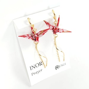 May include: A pair of gold-tone drop earrings featuring red and white paper crane charms with floral patterns. Each crane is suspended from a gold-tone hoop and attached to a delicate gold chain. The earrings are displayed on a white card with the text INOR Prayer and ENGI.