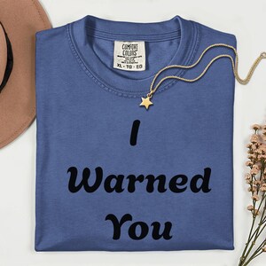May include: A blue short-sleeved t-shirt featuring the black text "I Warned You" printed on the front. The shirt has a Comfort Colors brand label at the inner neckline indicating size XL-TG-EG, and a gold-toned necklace with a star charm.