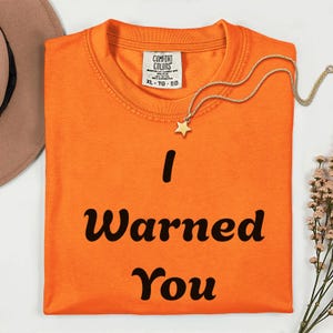 May include: An orange short-sleeved t-shirt with the black text I Warned You printed on the front. The shirt features a Comfort Colors brand label at the inner neckline, which indicates a size XL, and a gold-toned necklace with a star charm.