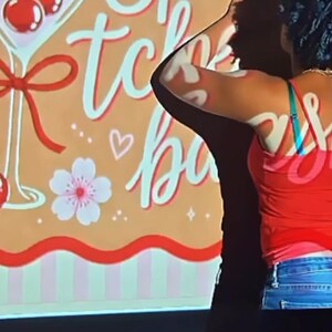 May include: A rectangular sign with a tan background featuring the text Spring tche bas in white script. The design includes a martini glass with three red cherries, a red ribbon, a white flower, and small white heart and star icons.
