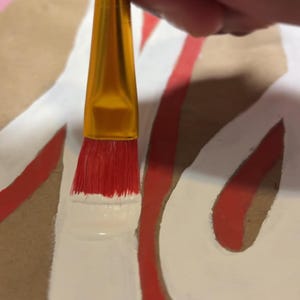 May include: A paintbrush with a gold-colored metal ferrule and a red handle is applying white paint onto a brown surface. The brush bristles are red and are currently coated in white paint, creating a thick, opaque white line on the brown material.