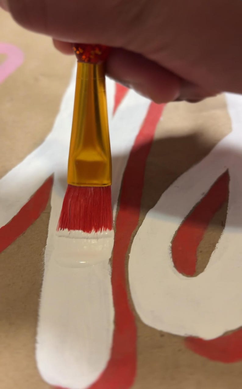 May include: A paintbrush with a gold-colored metal ferrule and a red handle is applying white paint onto a brown surface. The brush bristles are red and are currently coated in white paint, creating a thick, opaque white line on the brown material.