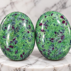May include: Two polished oval-shaped stones featuring a vibrant green base colour with scattered deep magenta and dark blue inclusions throughout the smooth, glossy surfaces of each individual stone, displayed side by side in a vertical orientation.