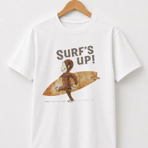 May include: A white short-sleeve t-shirt featuring a graphic of a brown monkey carrying a tan surfboard. The text SURF'S UP! is printed in a distressed dark brown font above the monkey. The design has a textured, weathered appearance.