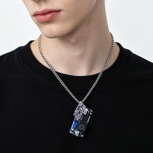 Op de afbeelding: Een zilverkleurige ketting met een rechthoekige hanger gemaakt van een blauwe en paarse elektronische printplaat met zichtbare componenten, waaronder kleine condensatoren en geïntegreerde chips, gedragen bij een zwart t-shirt.