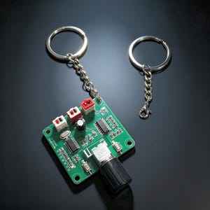 May include: A green square circuit board keychain featuring a black rotary knob, three small white and red connectors, and various electronic components. The board is attached to a silver metal split ring by a short silver chain, with an extra ring.